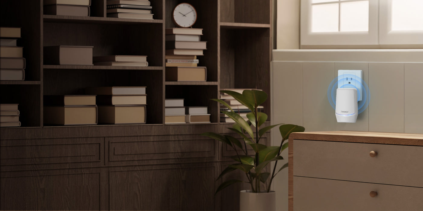 A white plug-in device with blue light rings indicating it's active, plugged into a wall outlet in a room with a wooden bookshelf full of books and a potted plant.