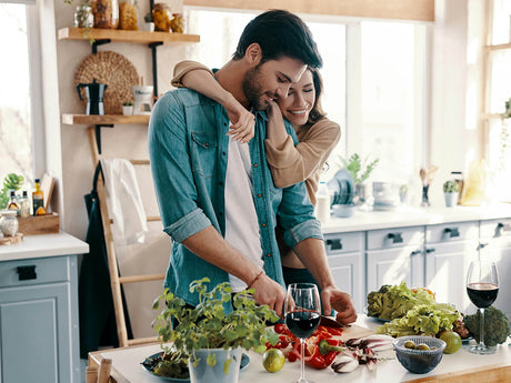 Paar bereitet gemeinsam eine Mahlzeit in der Küche zu, die Frau umarmt den Mann beim Gemüseschneiden.